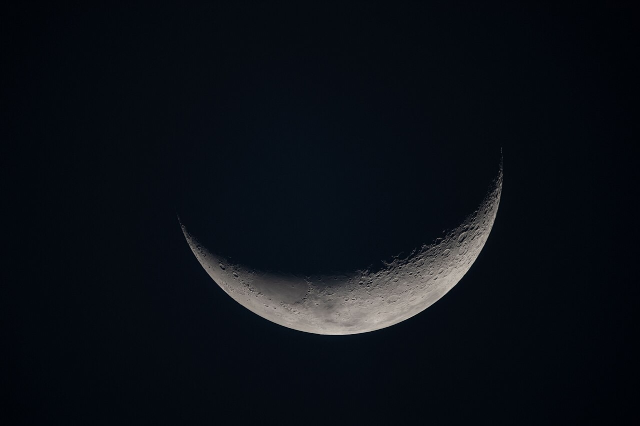 A thin sunlit lunar crescent with crater shadows along the terminator against black space.