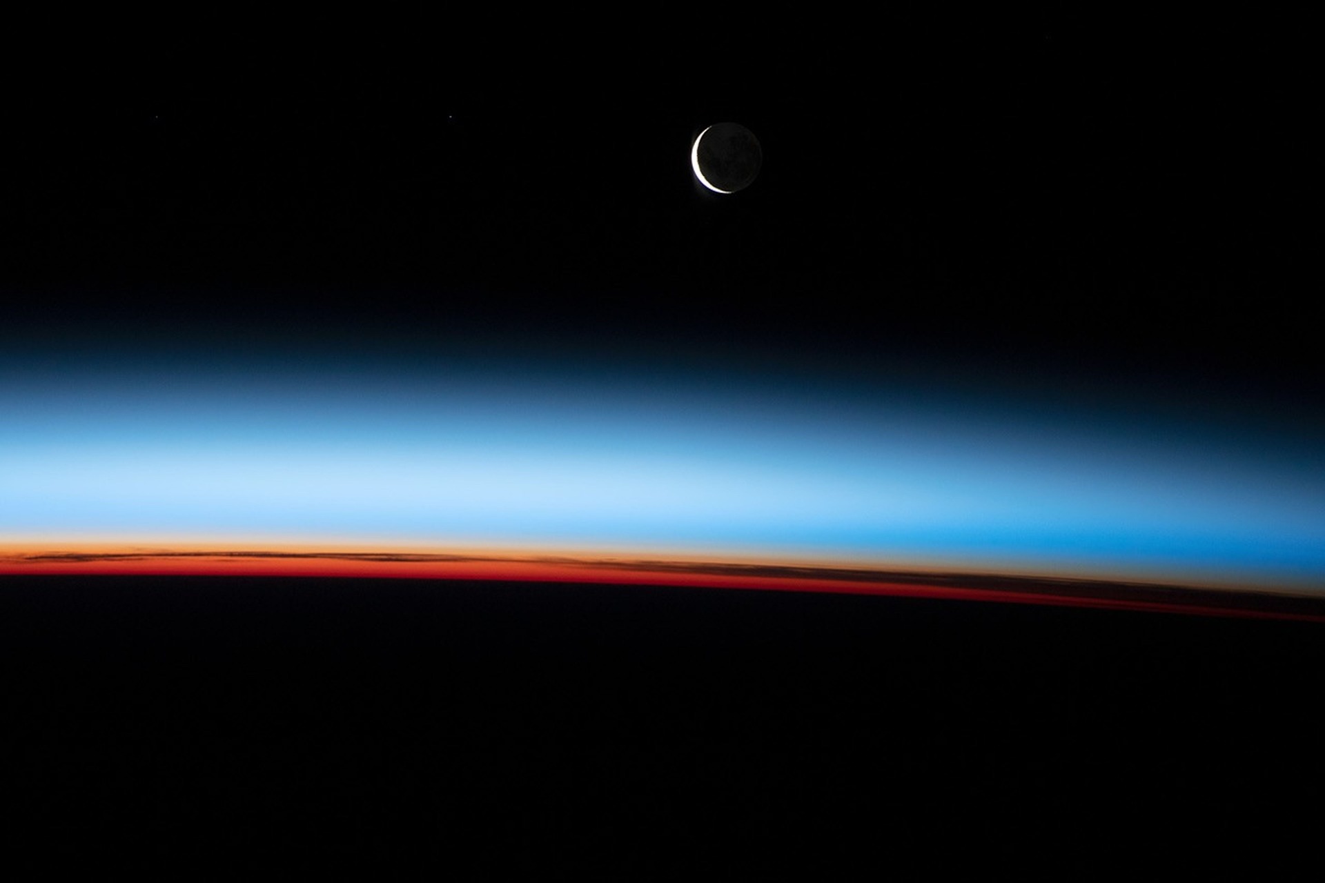 Orange and blue atmospheric layers above Earth's curved horizon, with a crescent Moon in the black sky.