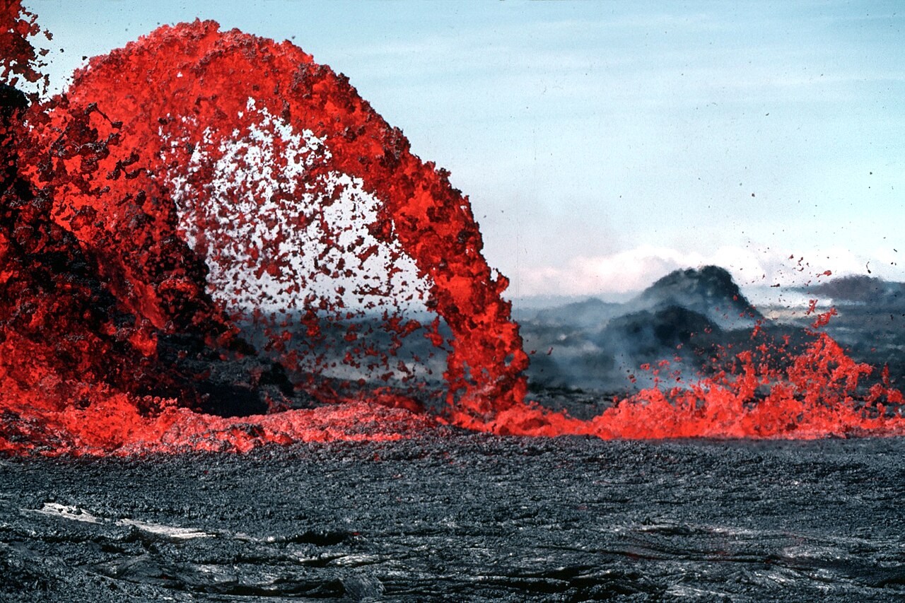 An arching orange lava fountain erupts from a vent on Kīlauea amid dark cooling lava.