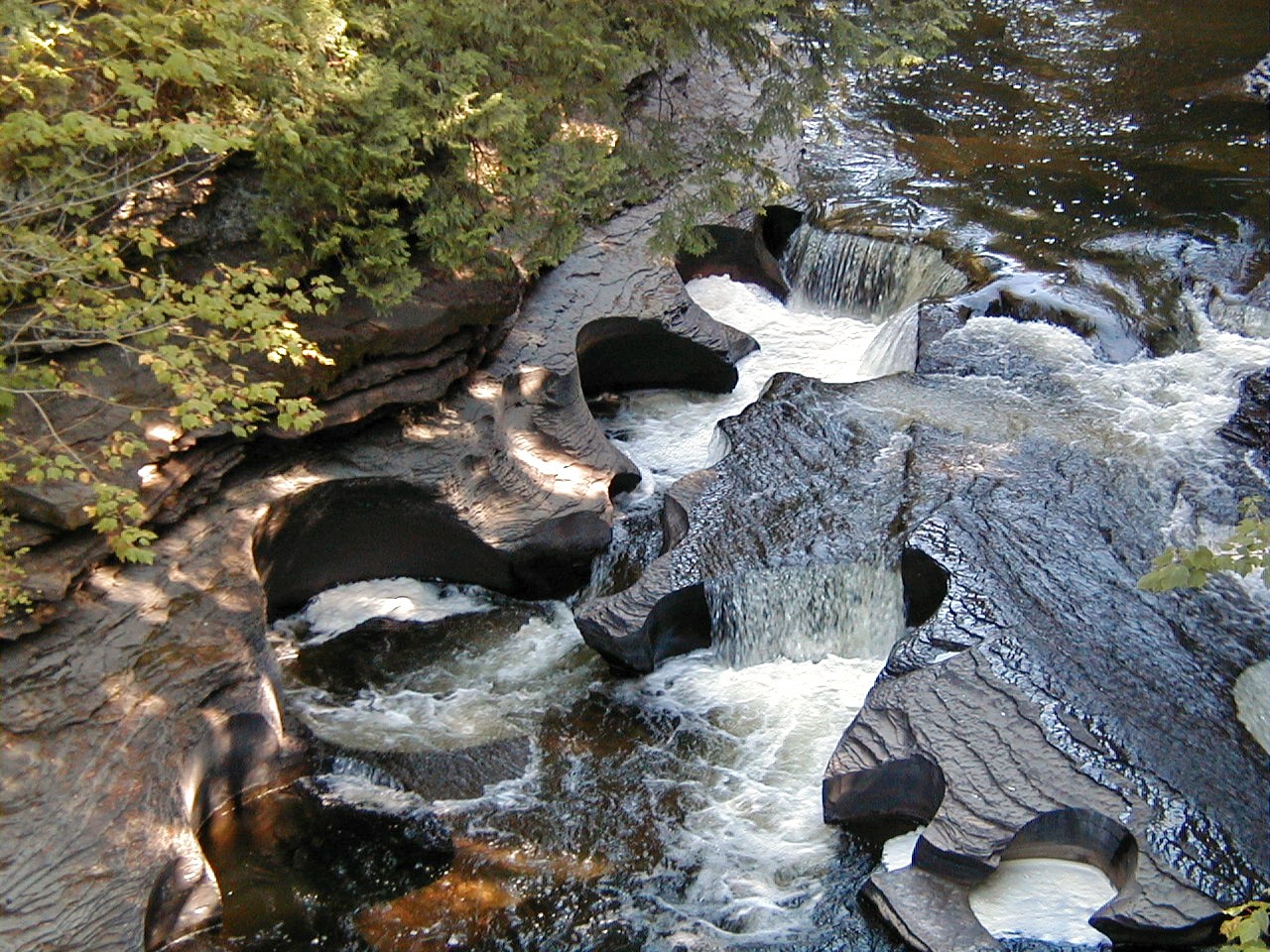 Rounded potholes cut into dark bedrock beside flowing water in the Presque Isle River, Michigan.